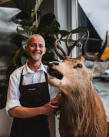 A man standing proudly next to a deer head trophy on the wall