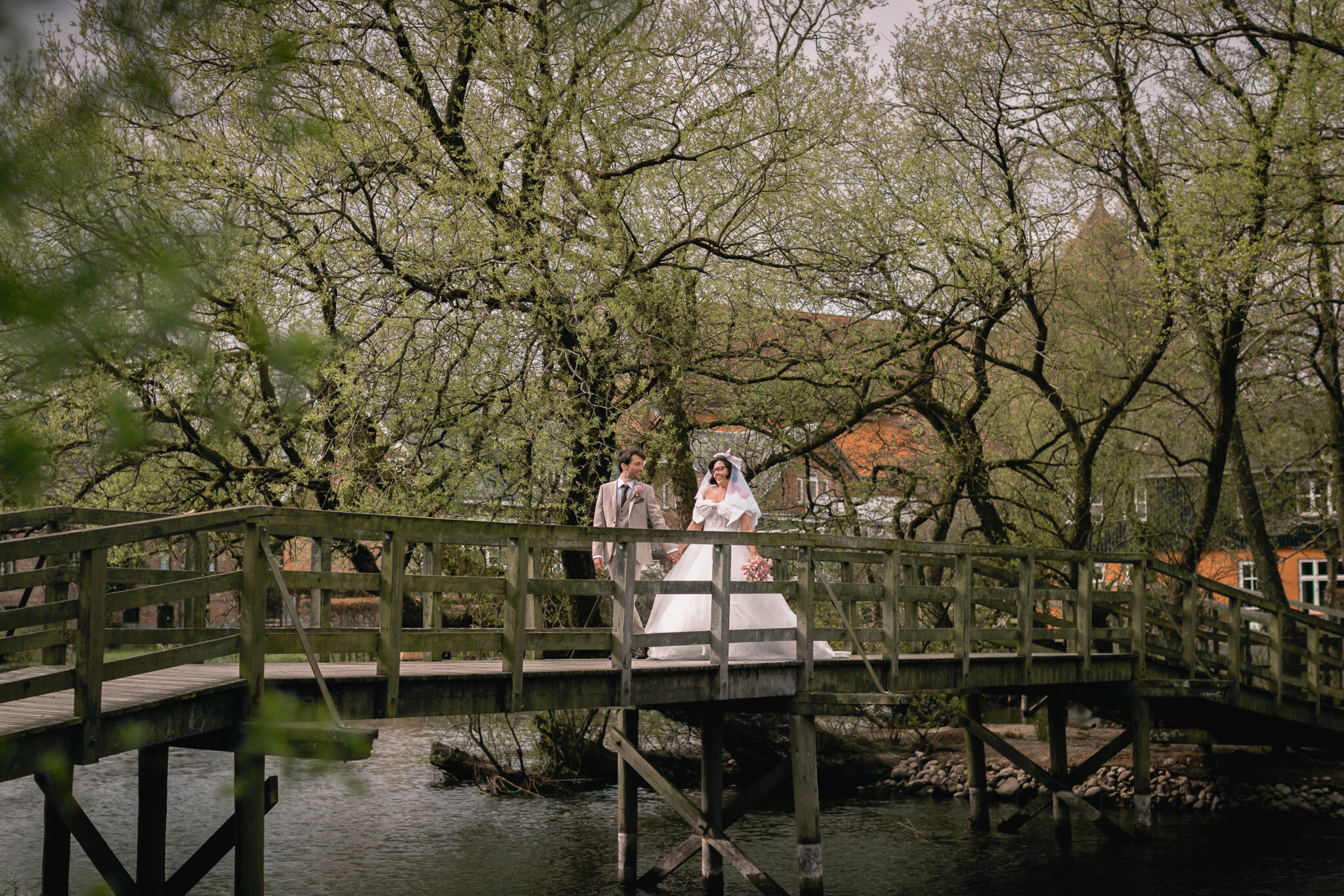A bride and groom stand on a wooden bridge surrounded by trees. The bride is in a white gown and veil, and the groom wears a light suit. It appears to be spring with lush greenery in the background—a perfect setting for exquisite wedding photography.