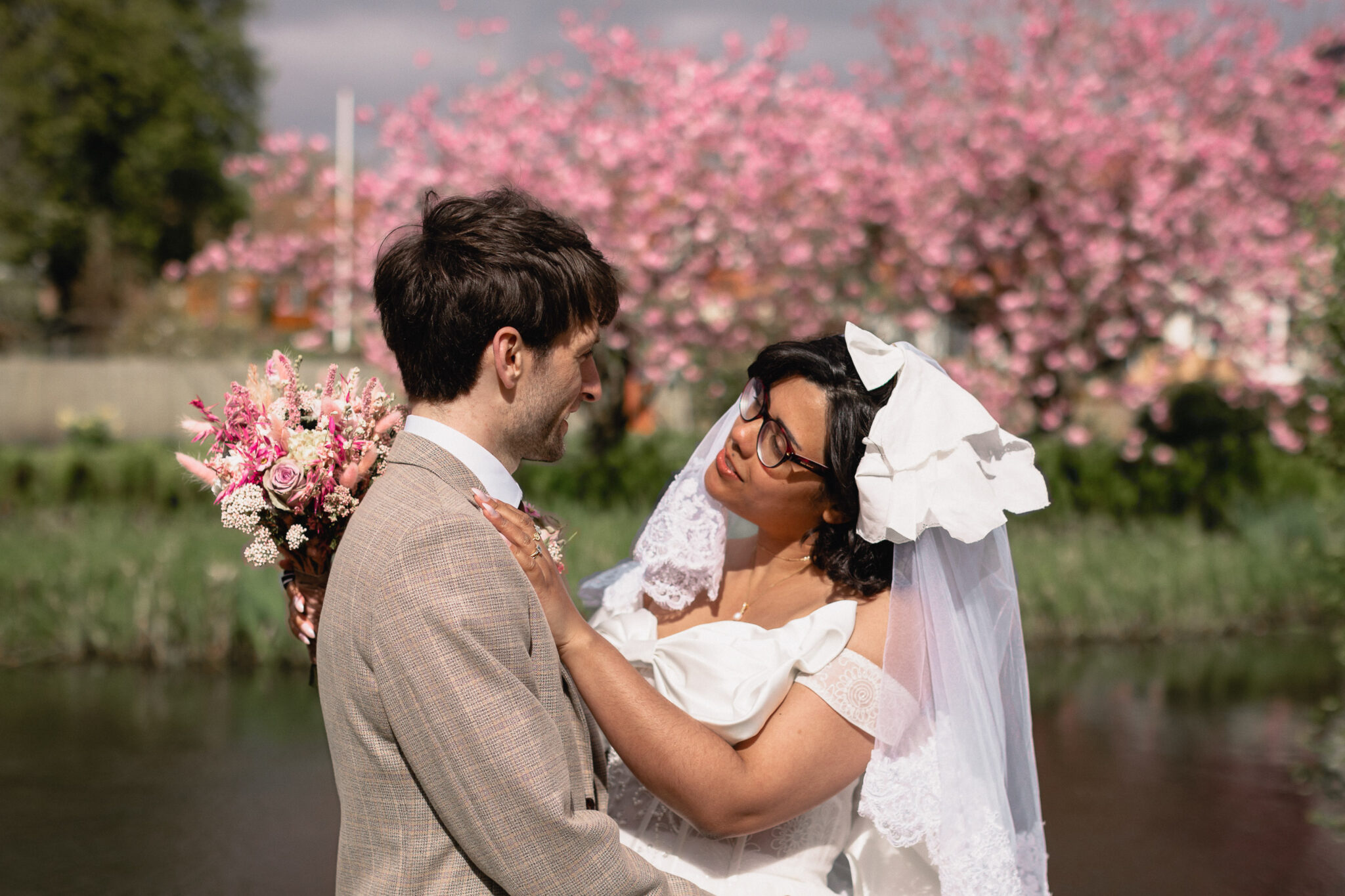 A bride and groom gaze at each other lovingly during their outdoor wedding ceremony, with blooming pink flowers in the background. Captured by exquisite wedding photography, the bride holds a bouquet, and both are dressed in traditional attire.