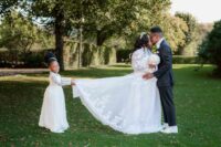 A bride and groom joyfully pose with their daughter in a serene park setting, surrounded by lush greenery