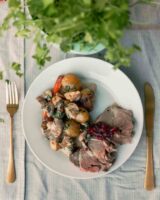 A plate of sliced roast meat with roasted potatoes, vegetables, and sauce, elegantly served with gold cutlery on a table