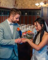 Couple exchanging rings during their wedding ceremony, with the groom in a light blue suit and the bride holding a bouquet