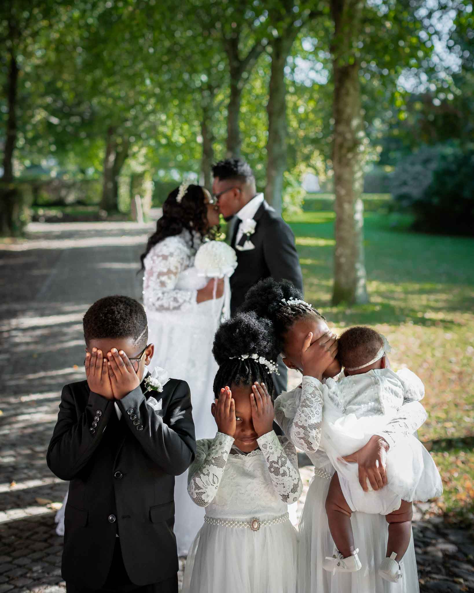 Charming wedding photo with children playfully covering eyes while bride and groom kiss in a beautiful park setting